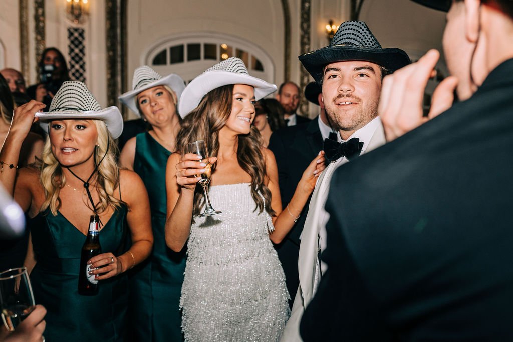 A group of people at a formal event, wearing cowboy hats and formal attire. A woman in a white dress holds a glass of champagne, while others are in dark dresses and suits, with some holding drinks. They appear to be indoors, possibly at a party or c