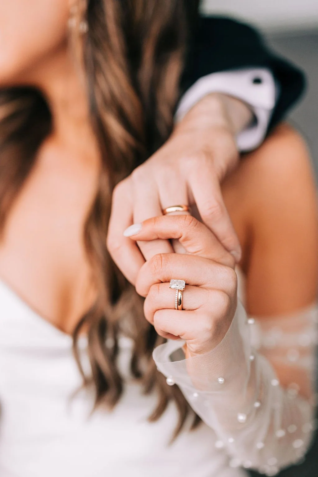 Close-up of intertwined hands of a couple, wearing wedding rings and engagement ring. The woman has a sheer sleeve with pearls, and the man is in a suit.