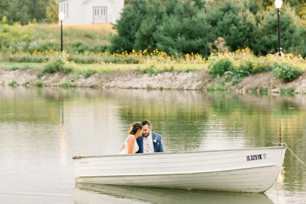 Couple sitting in a boat on a calm lake with trees and a building in the background.