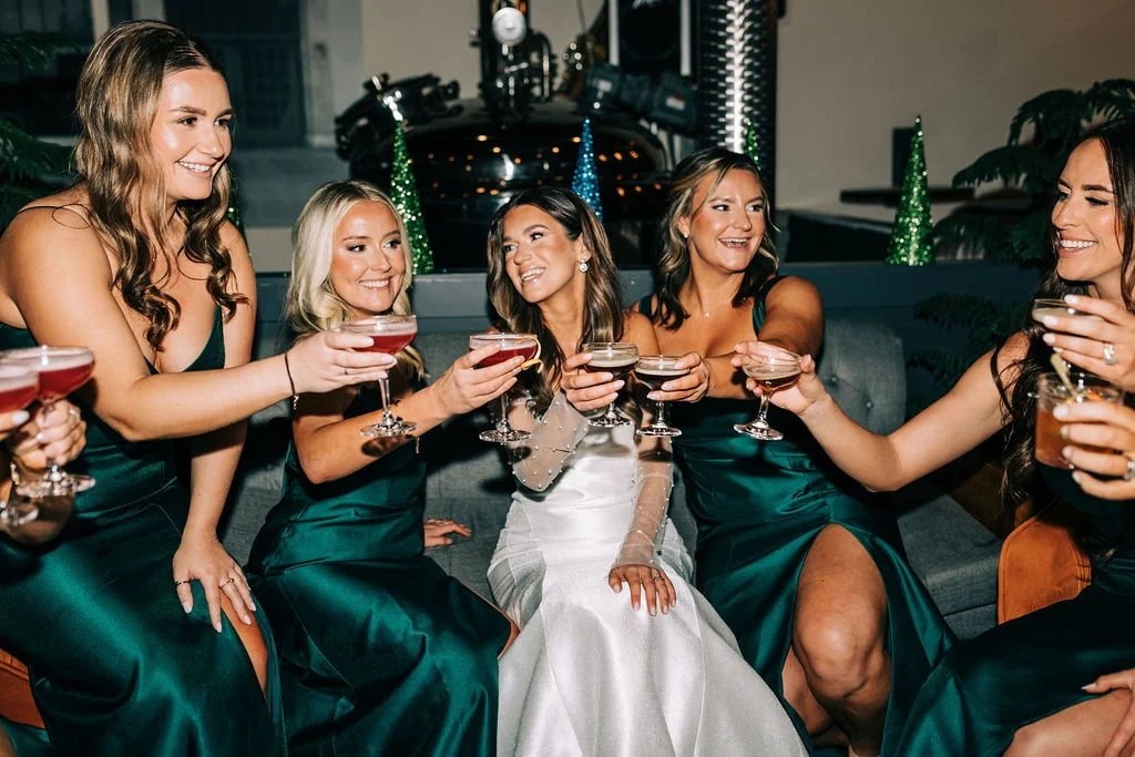 Bride and bridesmaids in green dresses toasting with cocktails indoors.