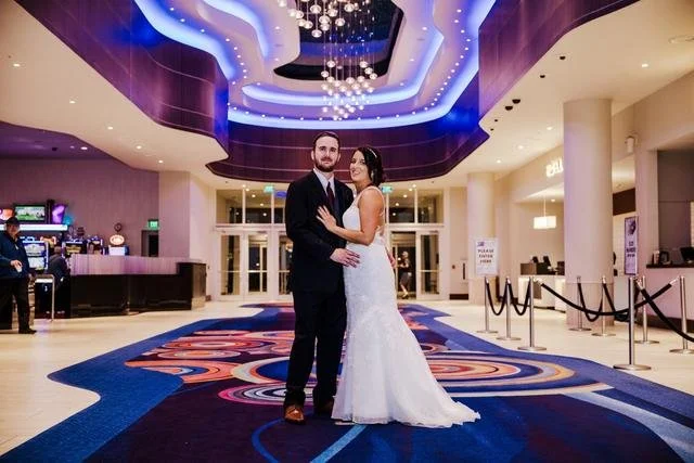 A couple dressed in formal attire is posing in a brightly lit hotel lobby with a modern design, featuring blue and purple ceiling lights and a colorful carpet.
