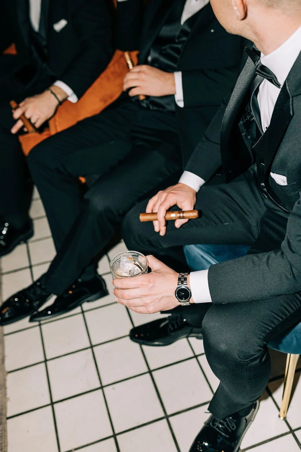 Men in formal attire holding cigars and drinks, sitting on a tiled floor.