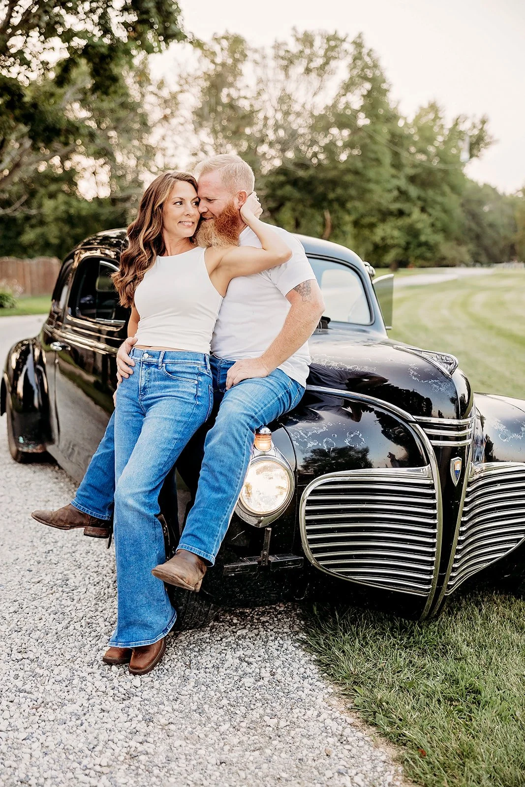 Couple sitting on vintage black car, embracing; both wearing casual outfits; outdoors with trees in the background.
