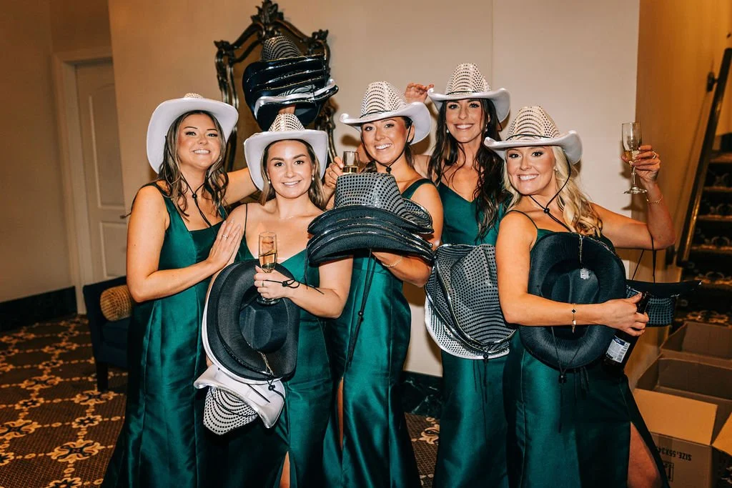 Five women in green dresses wearing cowboy hats, holding additional hats, and smiling indoors.