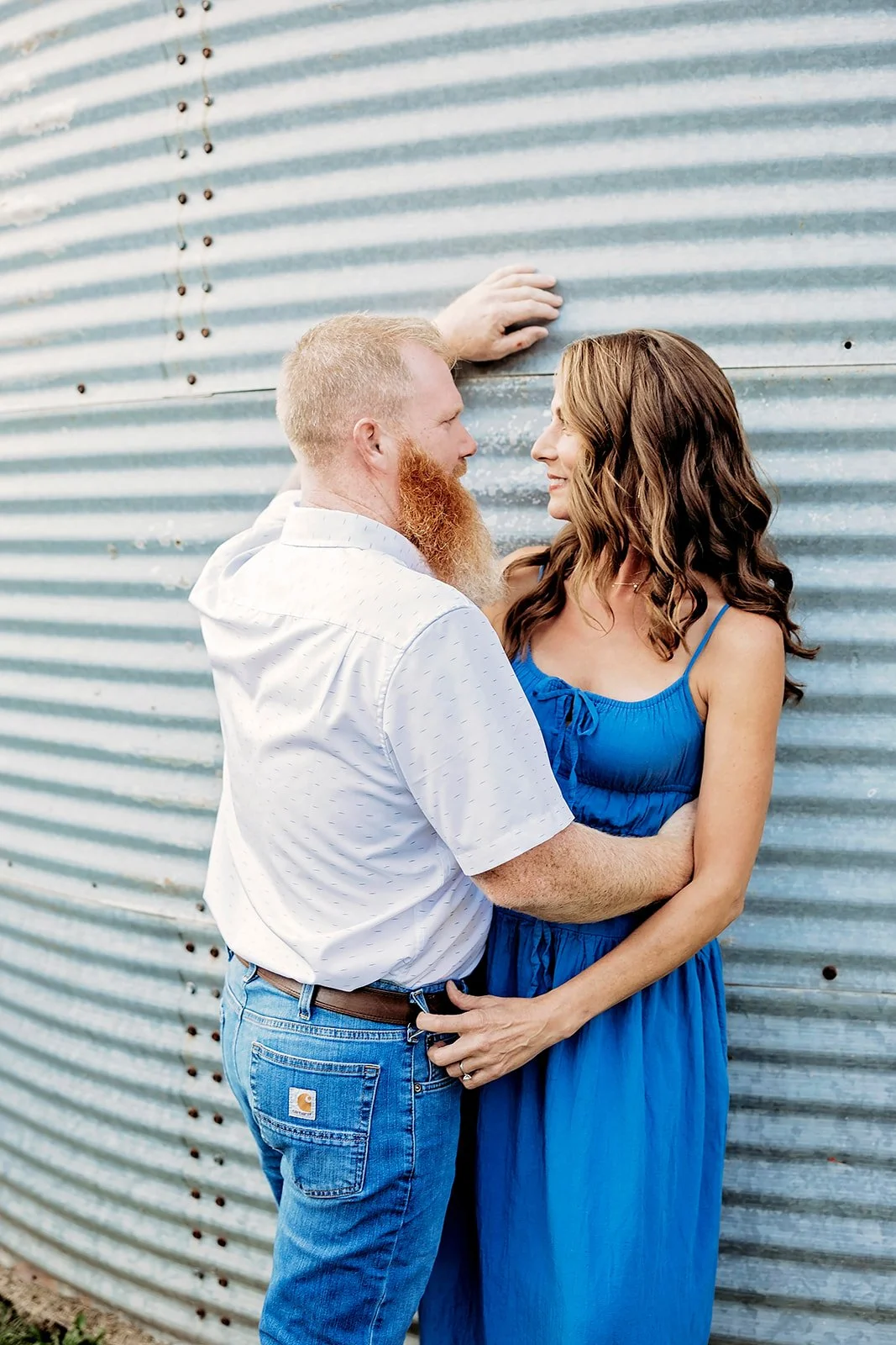 A couple standing closely in front of a corrugated metal wall. The man has a beard and is wearing a white shirt and jeans. The woman is wearing a blue dress.