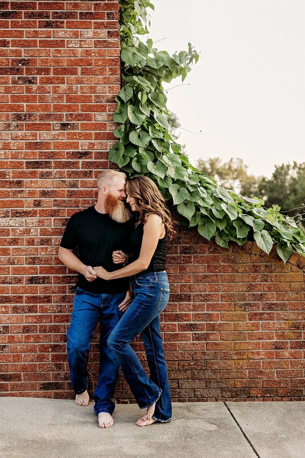 Couple affectionately leaning against a brick wall with vines.