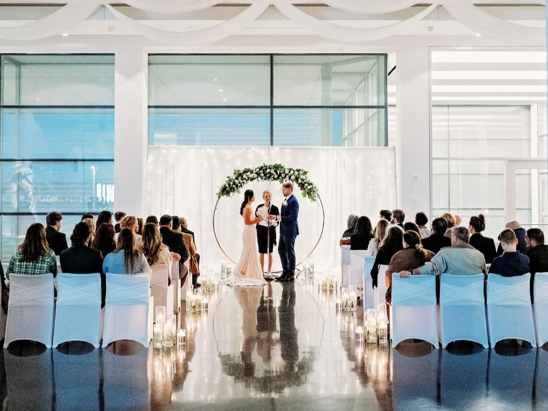 Indoor wedding ceremony with a bride and groom exchanging vows under a floral arch, surrounded by seated guests in a modern venue.