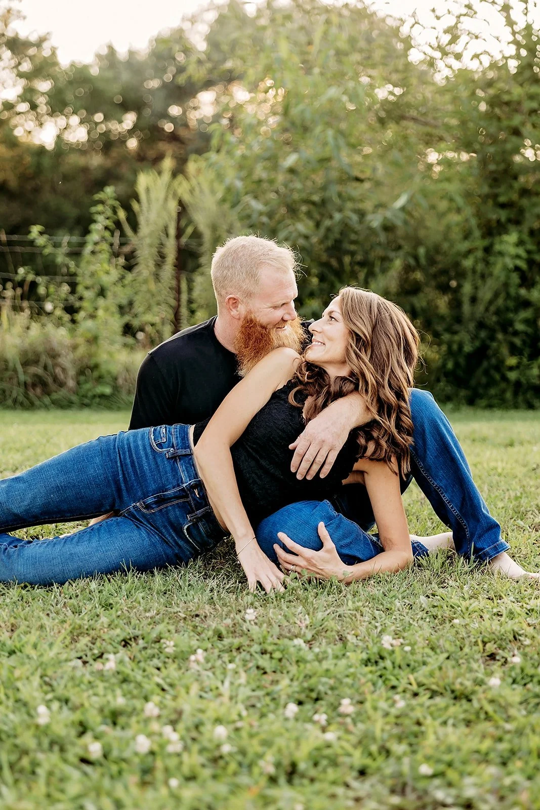 Couple sitting on grass, man with beard and woman smiling, outdoors with greenery in background.