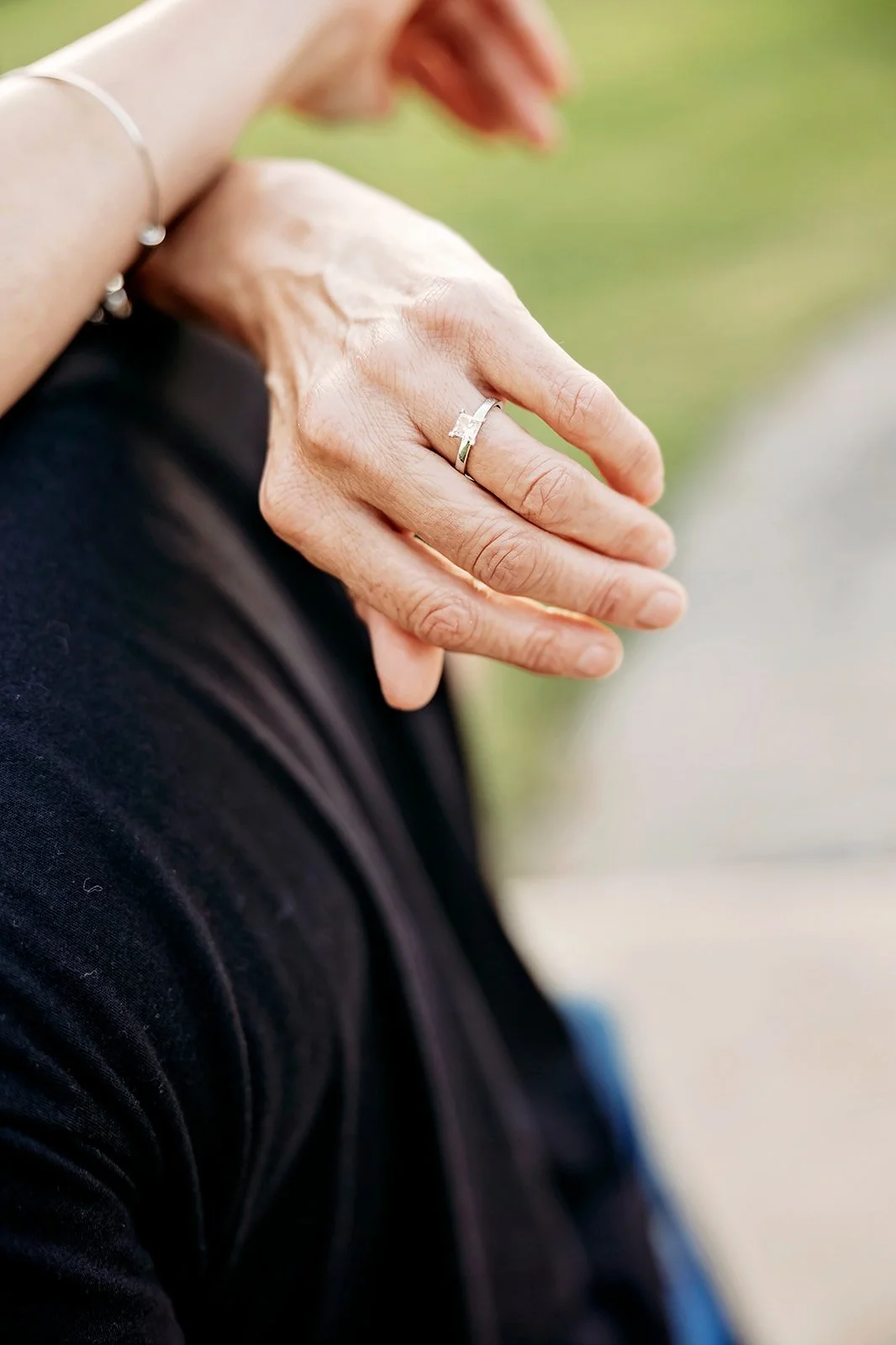 Close-up of a hand wearing a silver engagement ring with a square-cut diamond, resting on a dark fabric background.