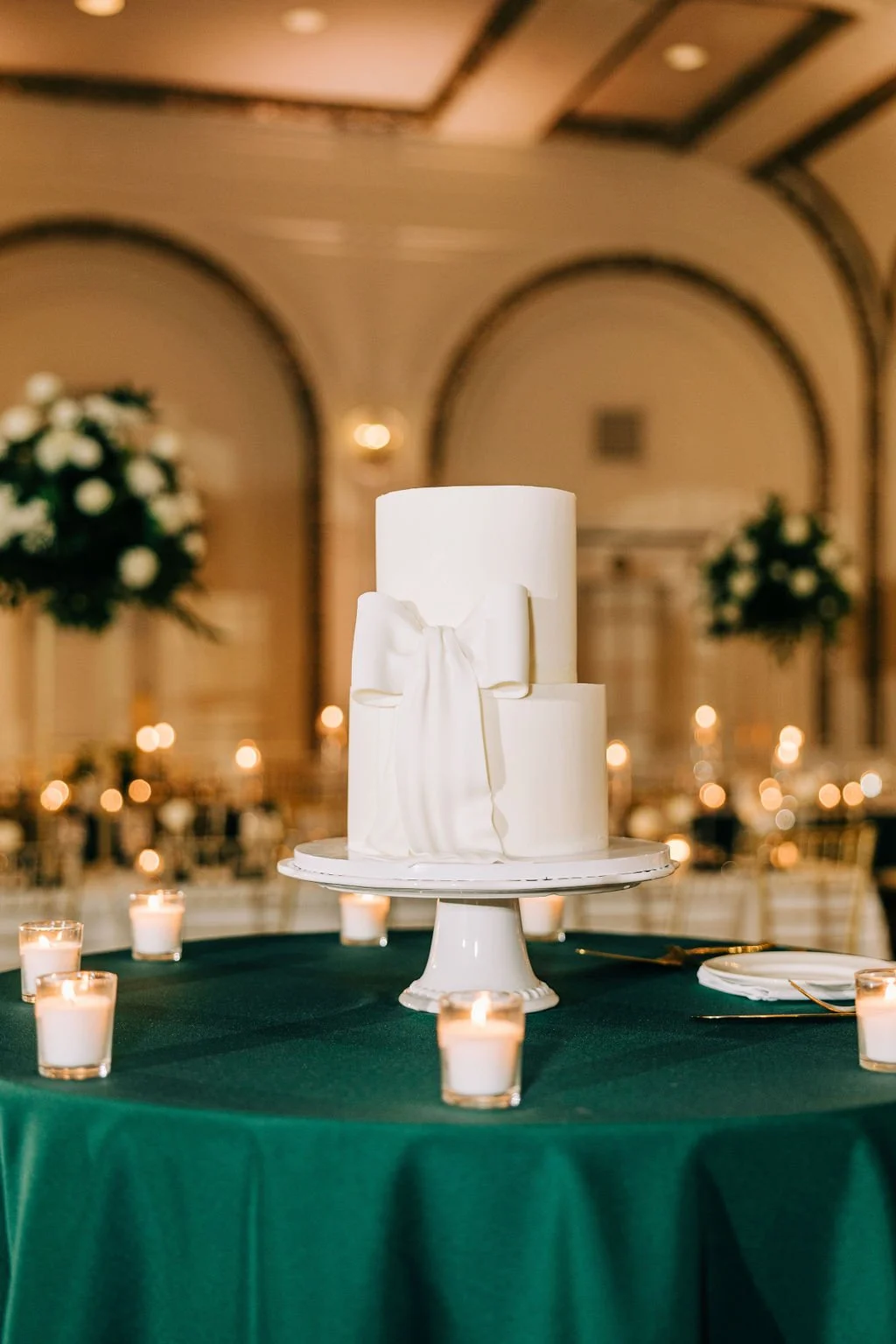 Elegant two-tier white wedding cake with bow decoration, surrounded by candles on a green table in a formal event setting.