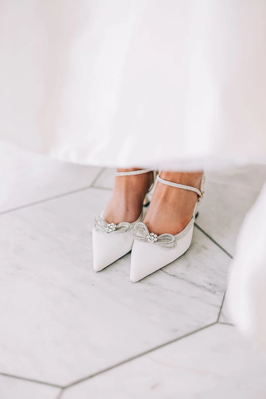 Close-up of a woman's feet wearing white pointed toe heels with decorative embellishments, on a white marble floor, partially covered by a white dress.