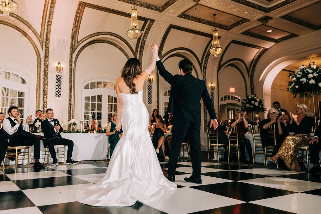Bride and groom dancing at a wedding reception in an elegant ballroom with guests seated and watching.