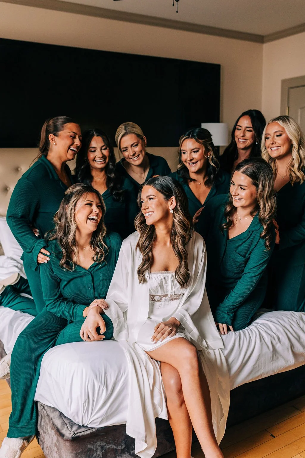 Group of women in matching green pajamas and a woman in a white robe sitting on a bed, smiling and interacting with each other in a cozy indoor setting.