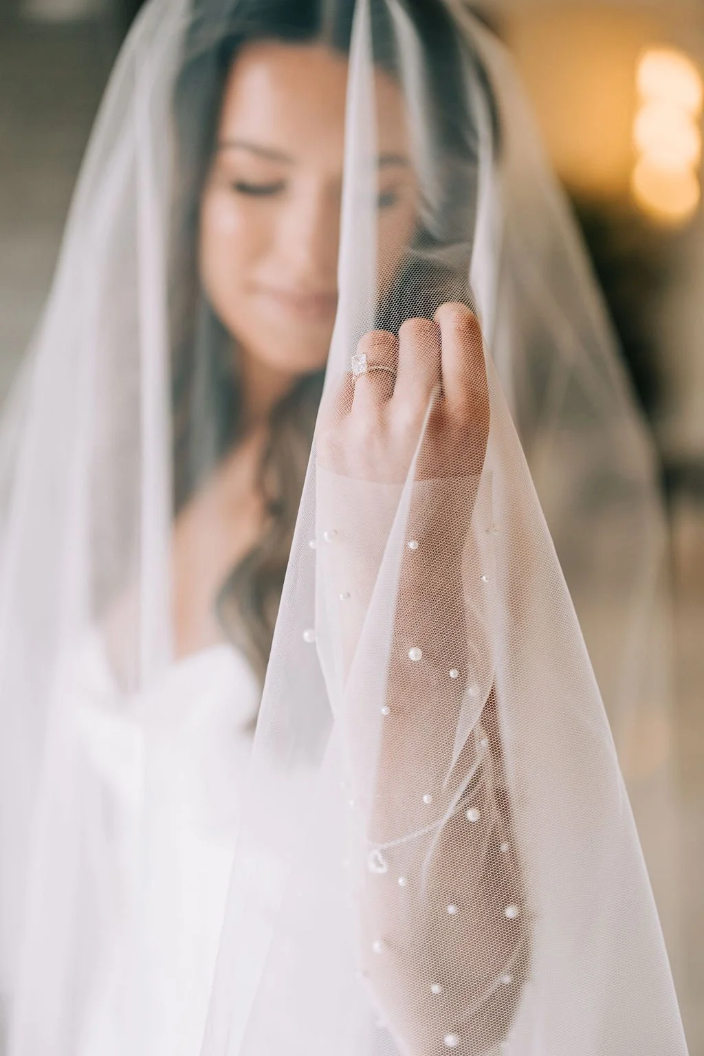 Close-up of a bride wearing a veil and holding it with her hand, featuring an engagement ring.