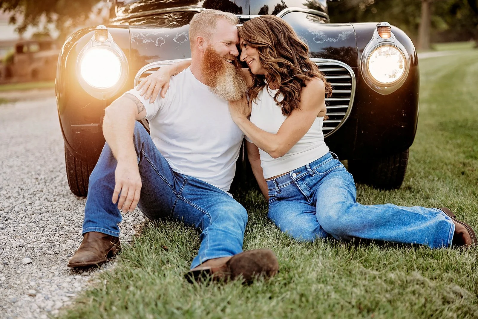 A couple sitting on the grass in front of a vintage car, both wearing jeans and white tops, with the car's headlights on.