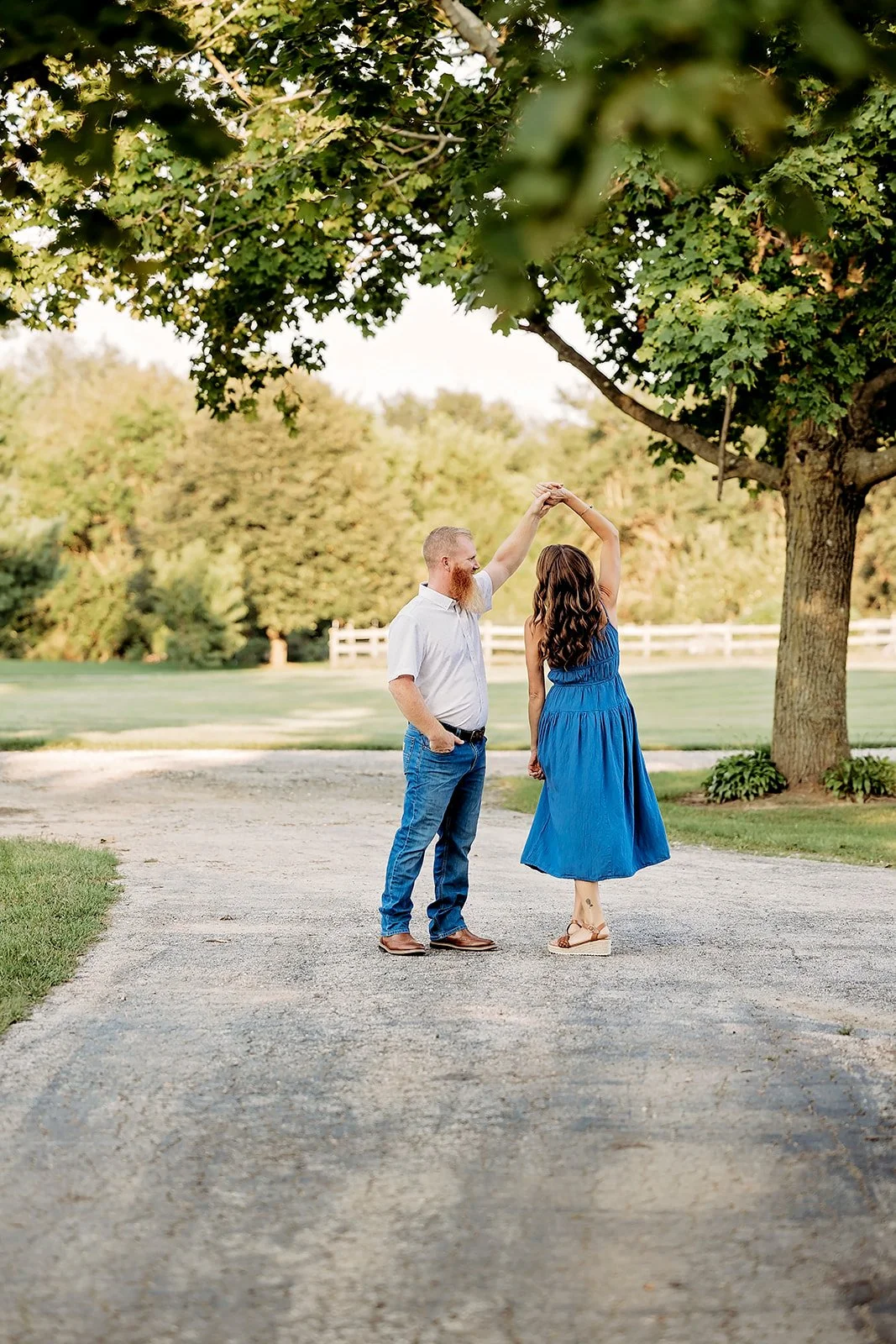 A couple dancing outdoors on a path under trees, with the woman in a blue dress and the man in jeans and a white shirt.