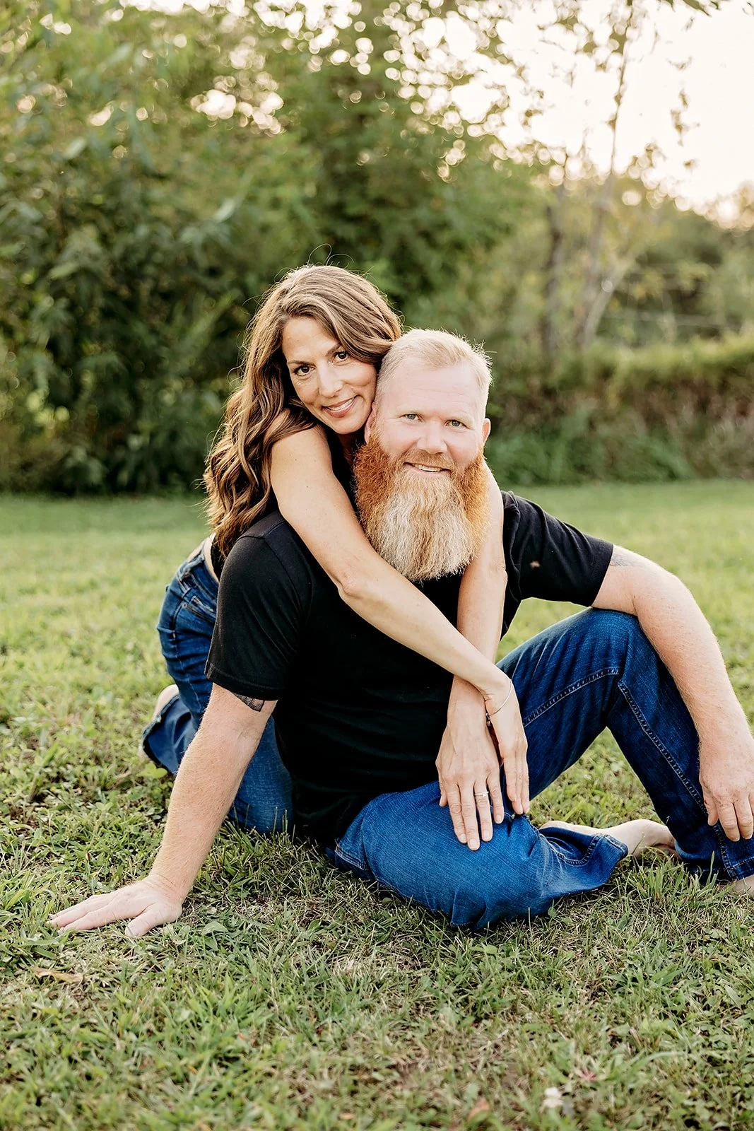 A woman is hugging a bearded man from behind as they sit on grassy ground, smiling at the camera in an outdoor setting.