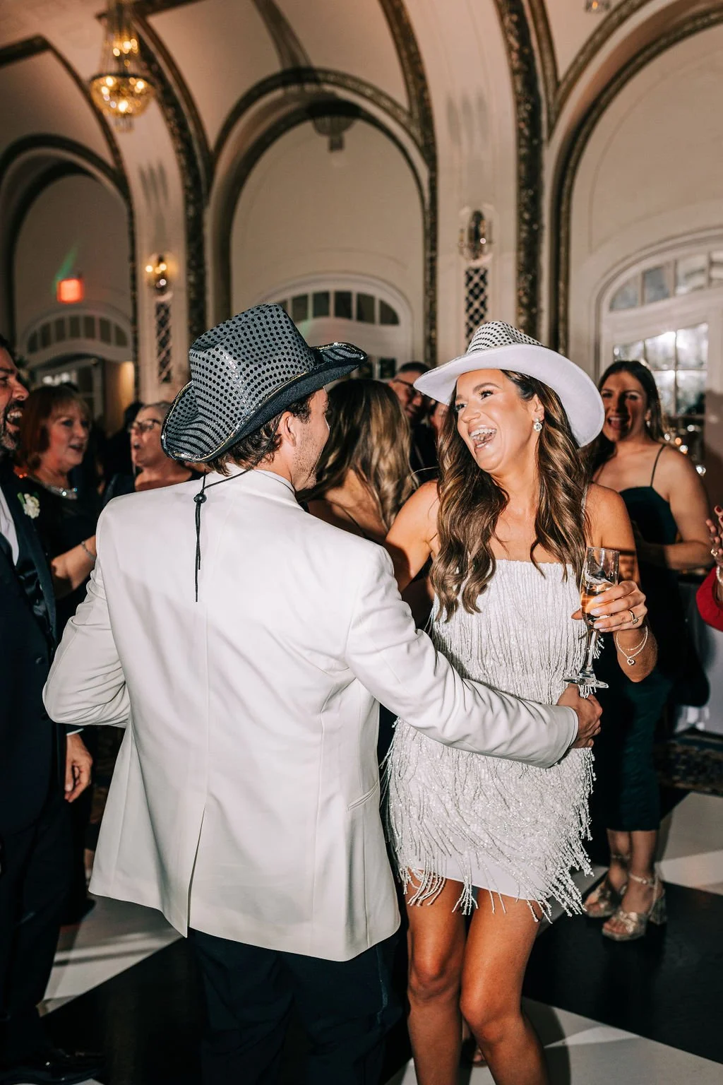 A couple dancing at an indoor party, both wearing cowboy hats. The man is in a white suit, and the woman wears a fringed dress and holds a drink. The background shows a decorated venue with other people enjoying the event.
