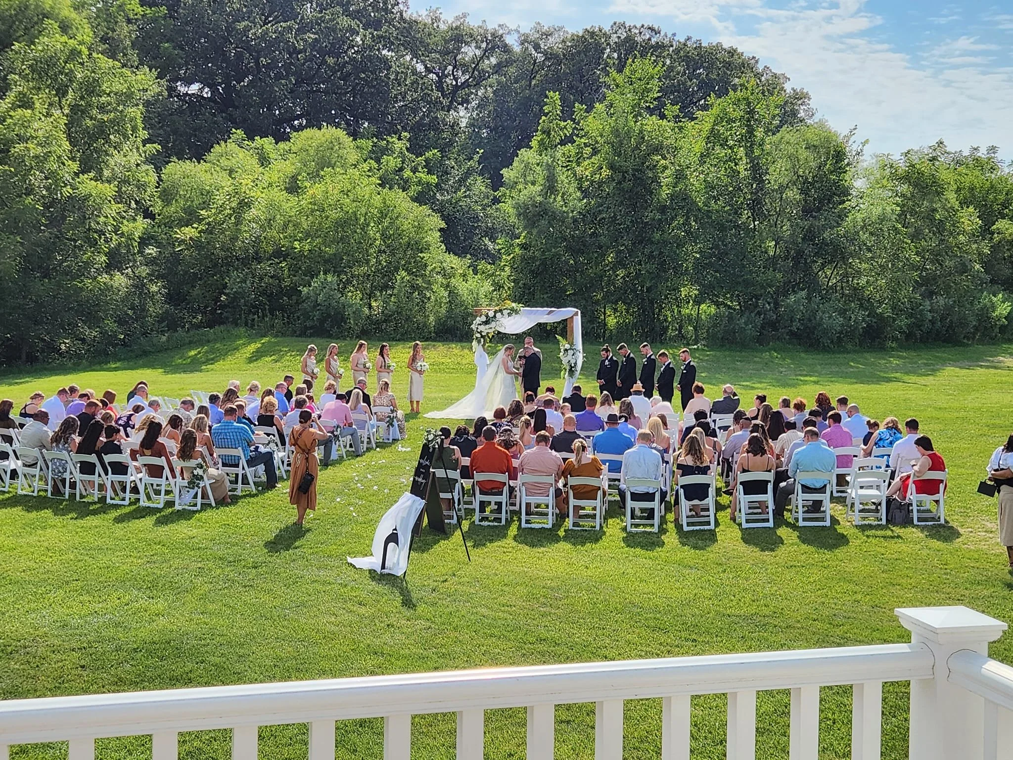 Outdoor wedding ceremony with guests seated on white chairs facing a decorated wooden arch, bridal party in formal attire, lush greenery background.