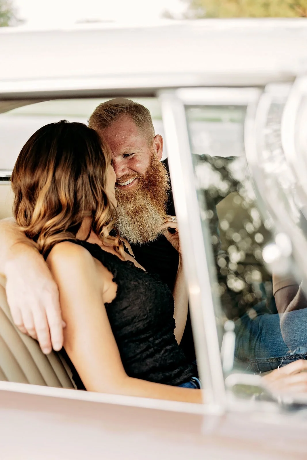 A couple sitting in a vintage car with the man smiling and leaning in towards the woman.