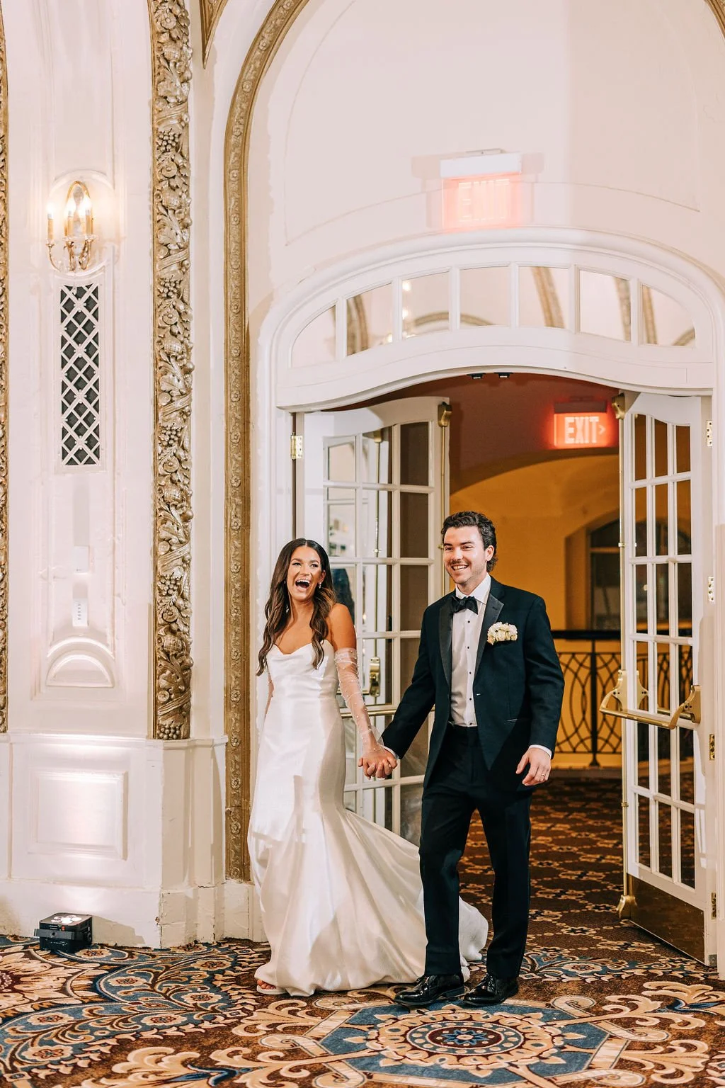 Bride and groom holding hands and smiling while entering a wedding venue, adorned with elegant architecture and ornate patterns on the carpet.