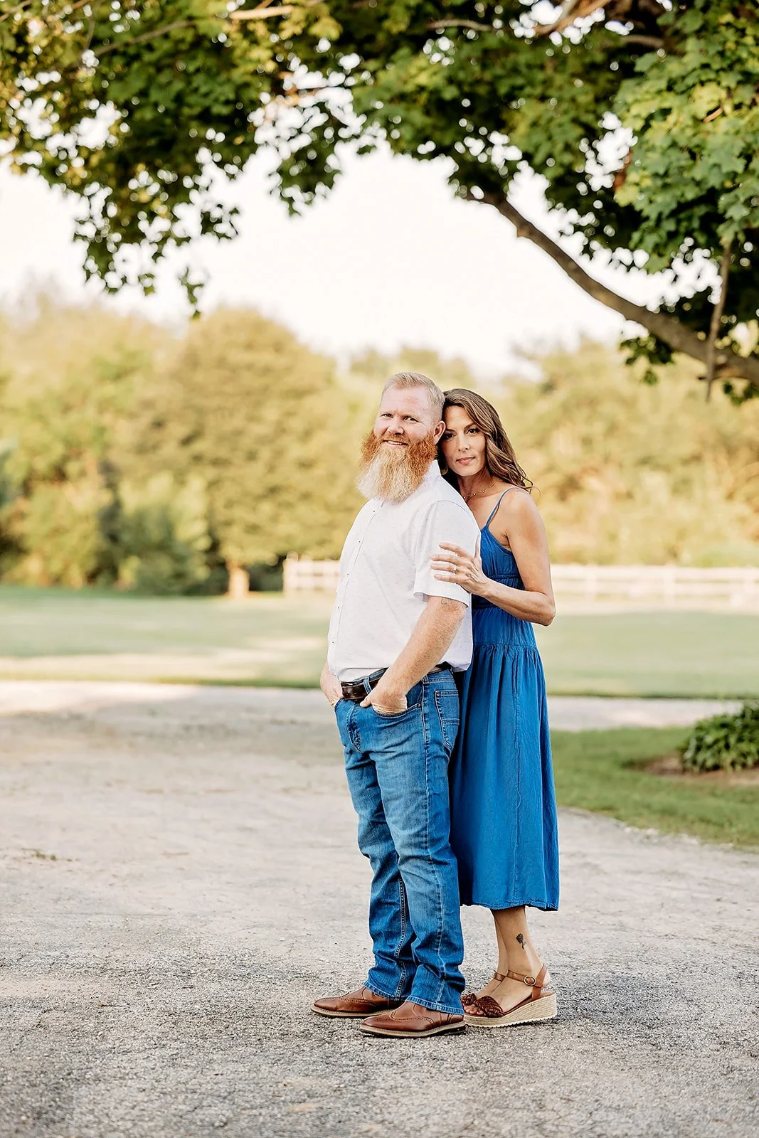 A man with a beard wearing a white shirt and jeans stands with a woman in a blue dress outdoors under a tree.