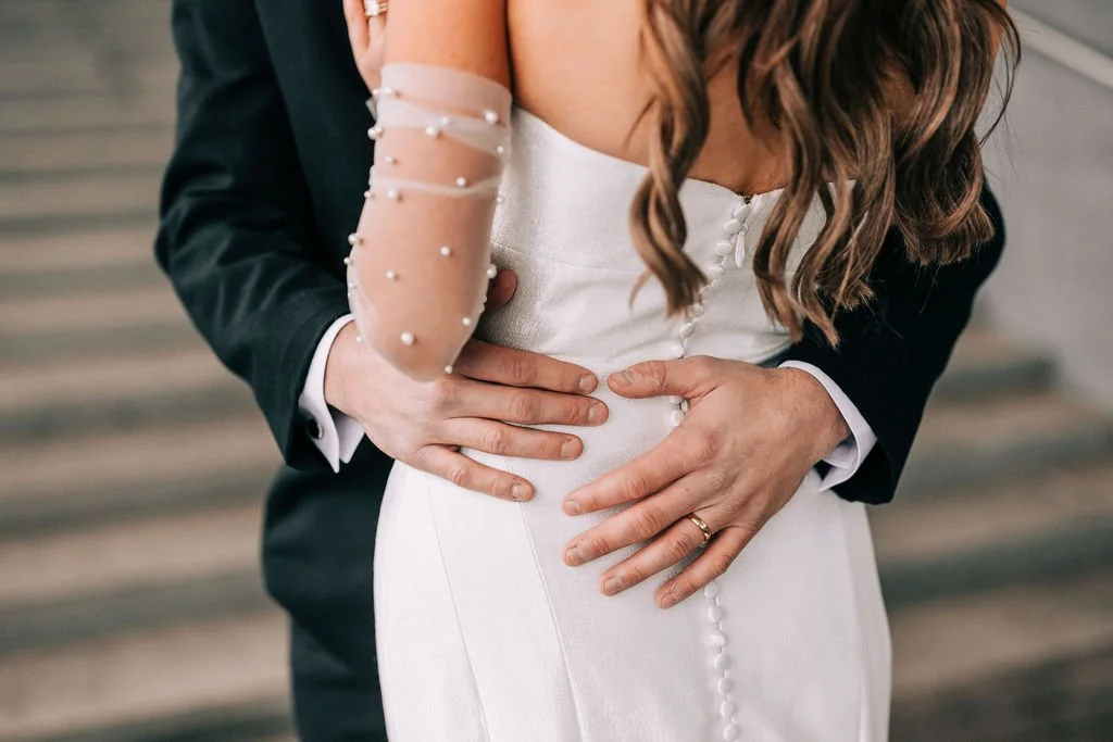 Bride and groom embrace, showing close-up of hands and wedding attire. Bride's white dress with button details and sheer sleeve. Groom's black suit and gold ring.