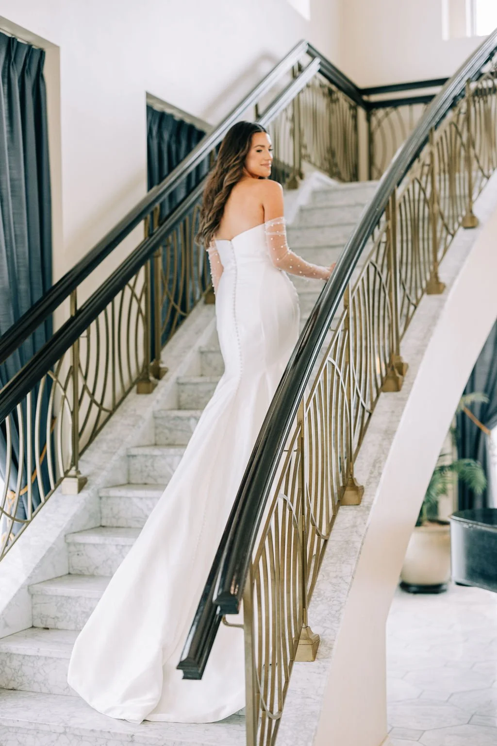 Woman in a white wedding dress ascending a staircase, with long hair and elegant design, featuring marble steps and stylish railings.