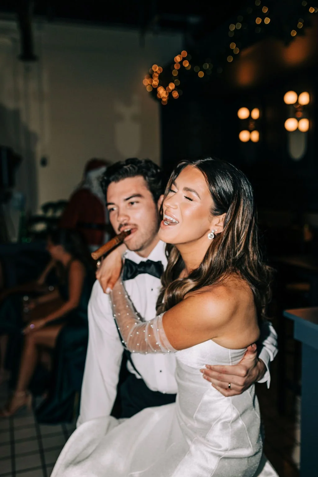 A couple, elegantly dressed, shares a joyful moment at a party. The woman is wearing a white dress and smiling, while the man, in a suit with a bow tie, holds a cigar. The setting includes dim lighting and decorative lights in the background.
