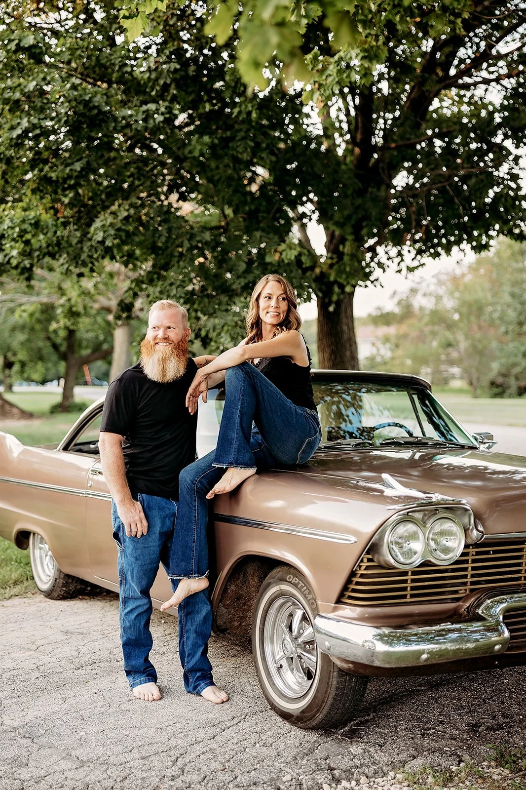 Man with a beard standing by a vintage car, a woman sitting on the car hood, both barefoot, in a leafy outdoor setting.