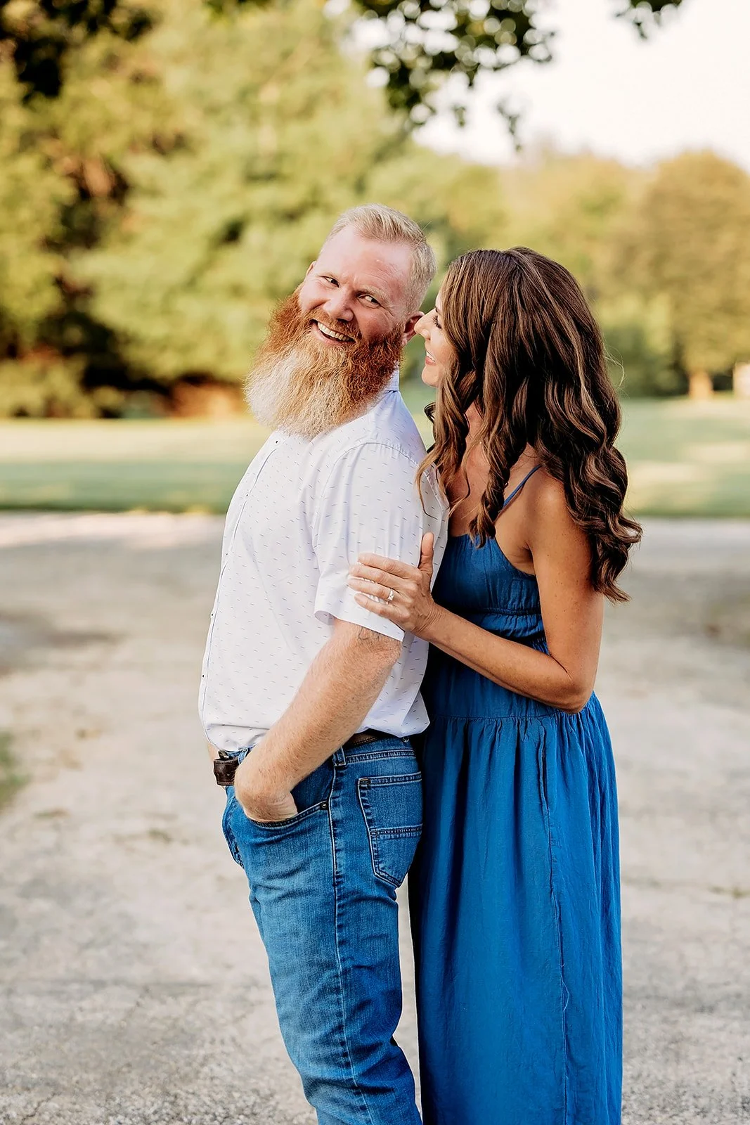 A smiling couple standing close together outdoors, with the man wearing a white shirt and blue jeans, and the woman in a blue dress, gently holding his arm.