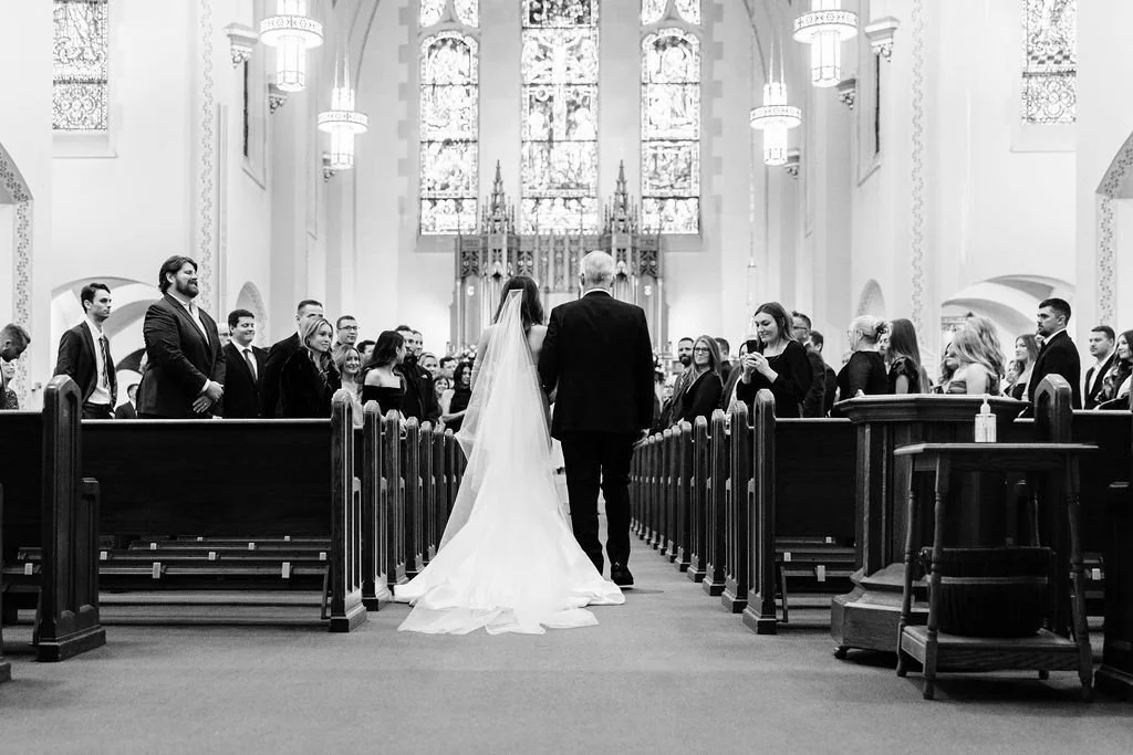 Bride and father walking down the aisle in a church during a wedding ceremony, with guests seated in pews and stained glass windows in the background.