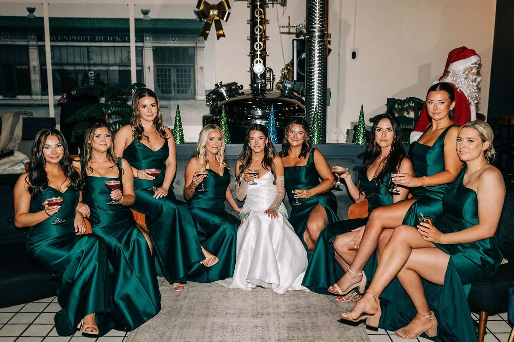 A bride in a white dress seated with bridesmaids in matching green dresses, holding drinks, indoors with festive decorations.