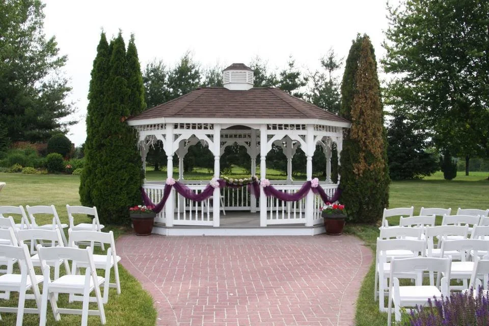 Outdoor wedding setup with white wooden gazebo, purple drapery decorations, surrounded by white folding chairs on a brick path, and greenery in the background.