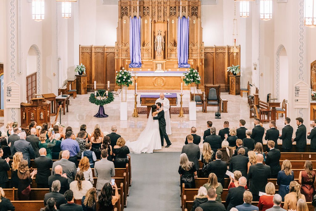 A wedding ceremony in a church with a couple sharing their first kiss. The bride and groom are at the altar, surrounded by a decorative setup with flowers and a backdrop featuring wooden details. Guests are seated, dressed in formal attire, witnessin