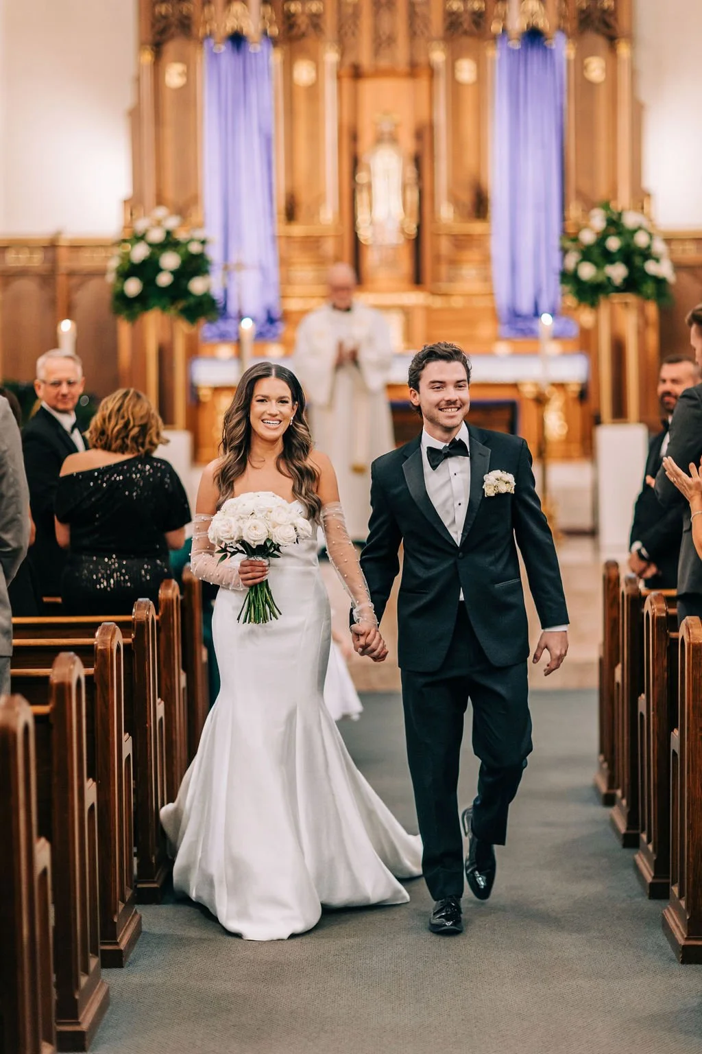 Bride and groom walking down a church aisle, smiling, with the bride holding a bouquet of white roses. They are wearing a wedding dress and a tuxedo. The church is decorated with flowers and an altar in the background.