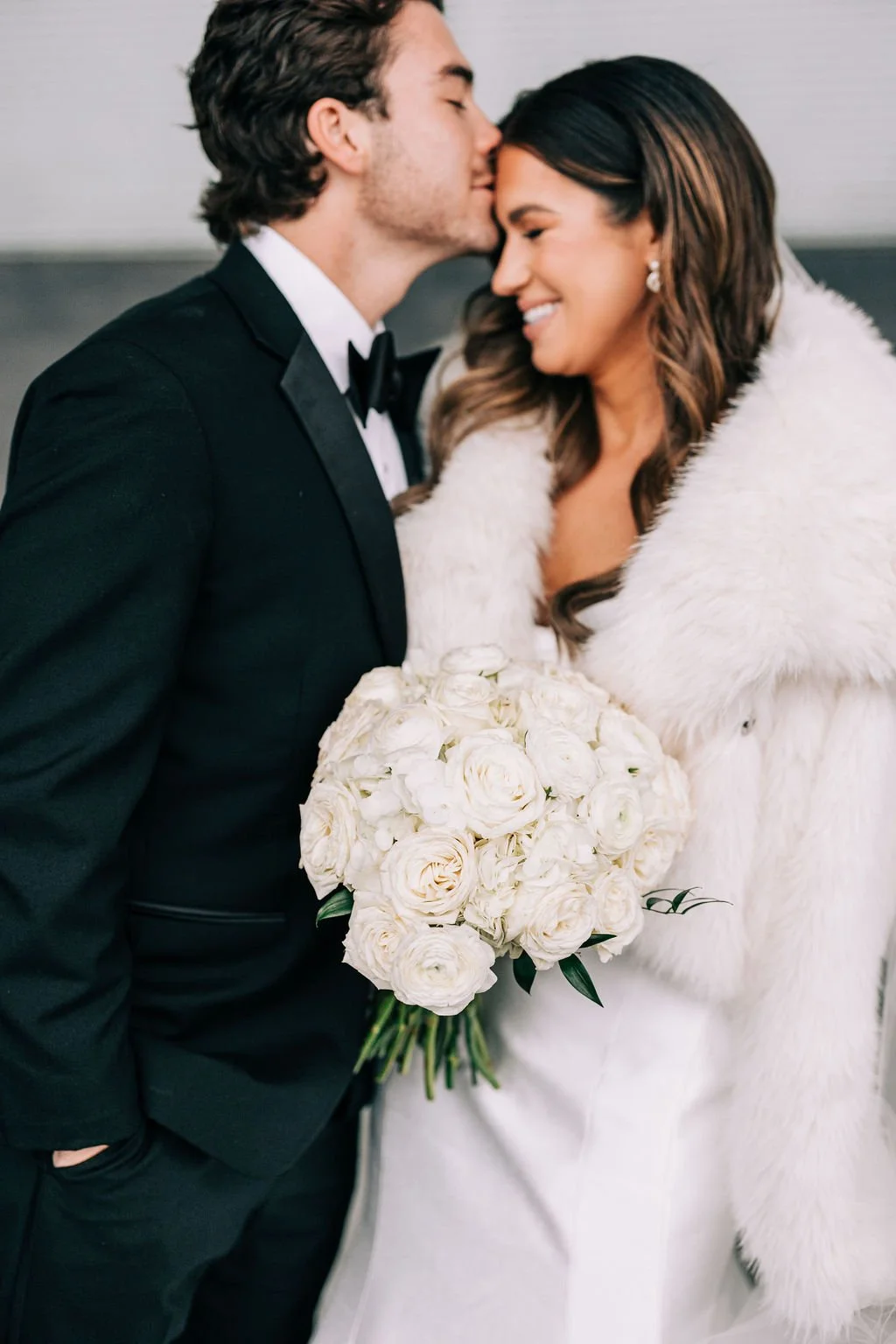 Bride and groom embracing, holding a bouquet of white flowers, groom in black tuxedo, bride in fur coat.
