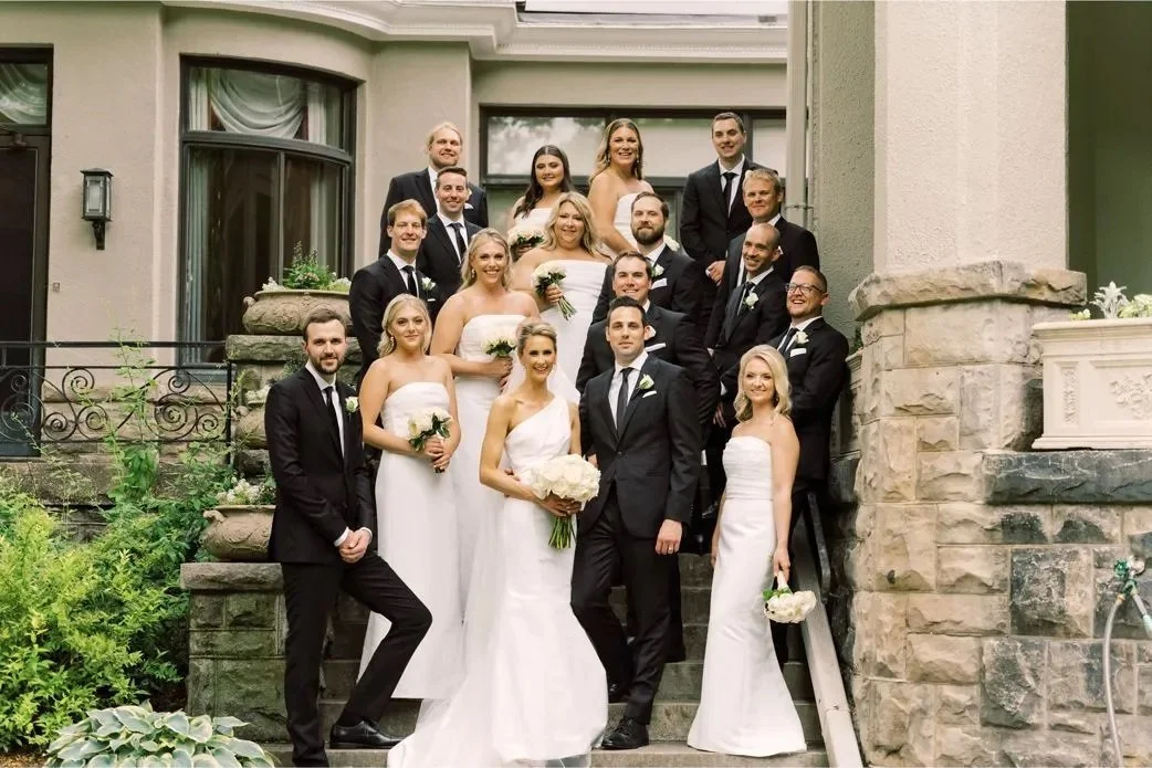 A wedding party group photo on steps with bridesmaids in white dresses and groomsmen in black suits.