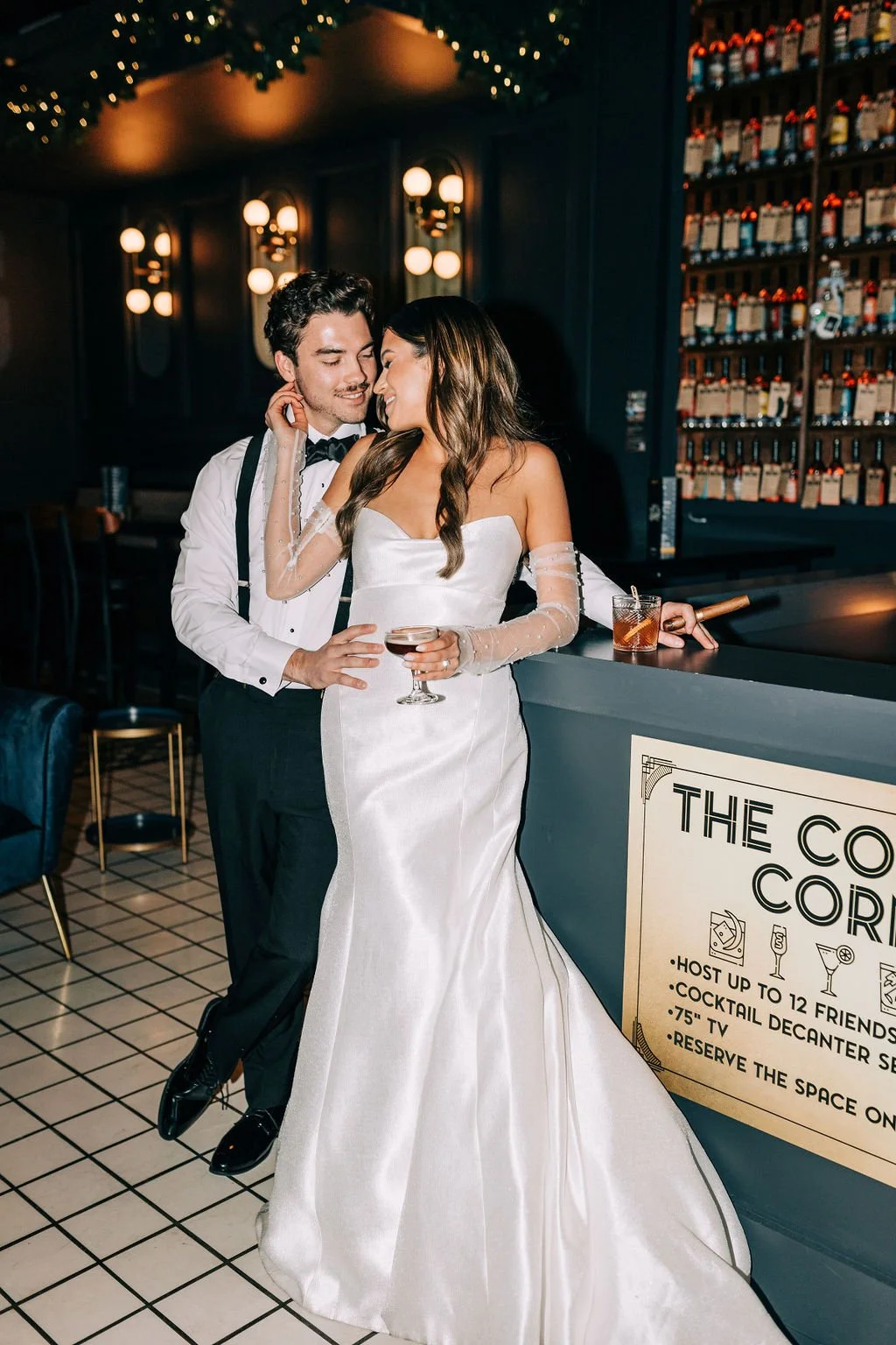 A smiling couple in formal attire, with the woman in a white wedding dress holding a cocktail and the man in a tuxedo, embracing at a bar with a "The Cocktail Corner" sign.