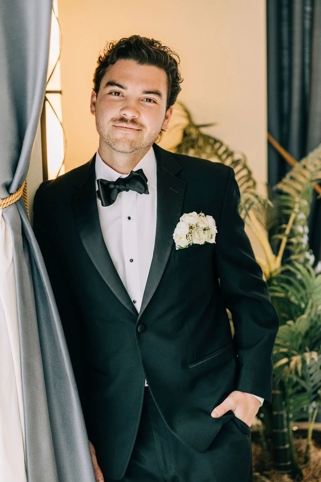 Man in a tuxedo with floral boutonniere, standing next to a curtain and plants.