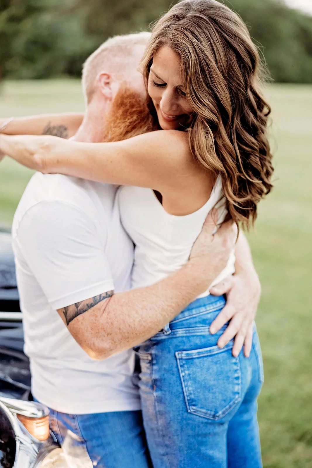 A couple embracing outdoors near a car, with the woman smiling and wearing a white top and jeans.