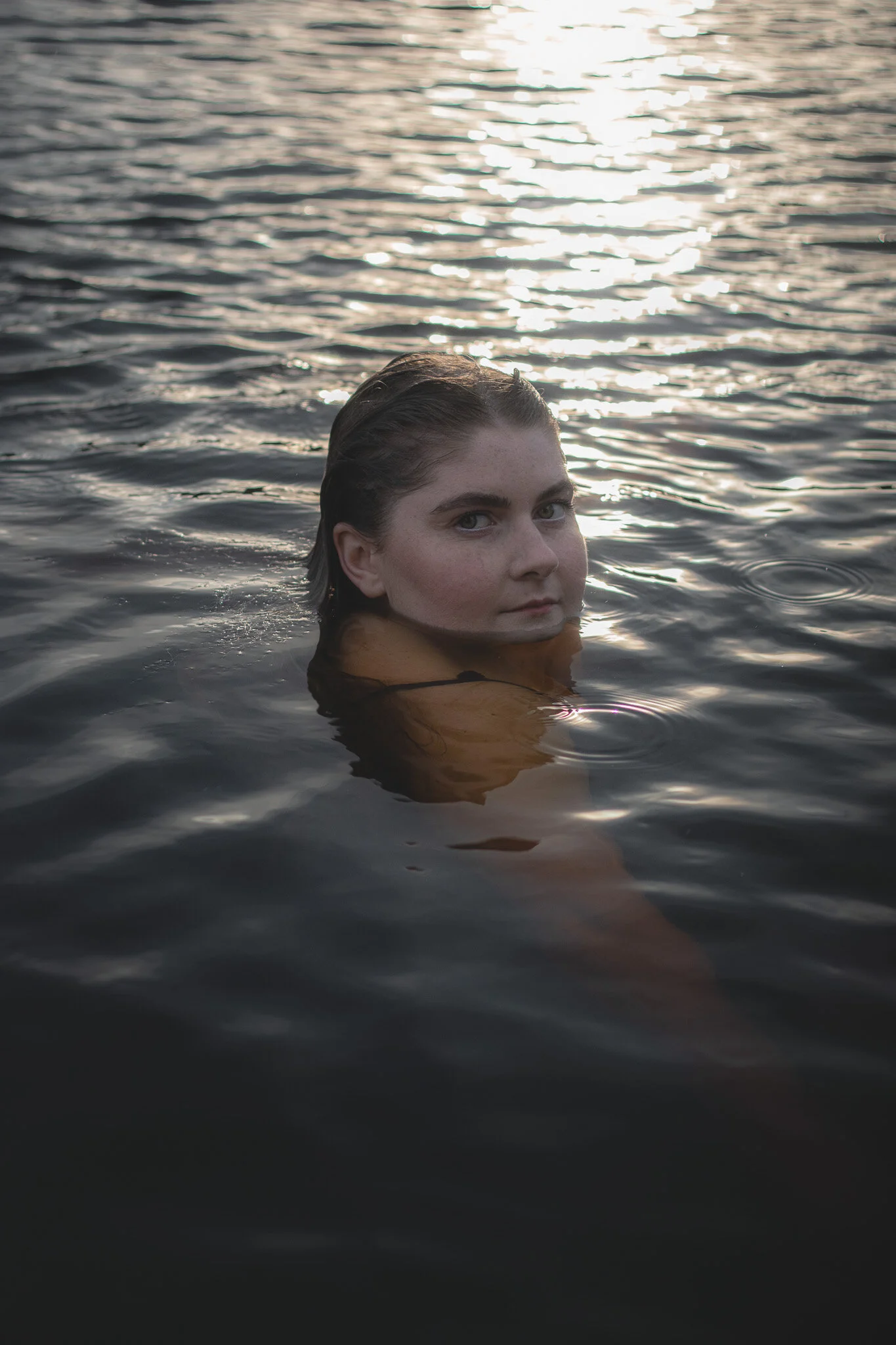 A woman with wet hair floating in dark water with a reflective surface, seen during sunset or sunrise.