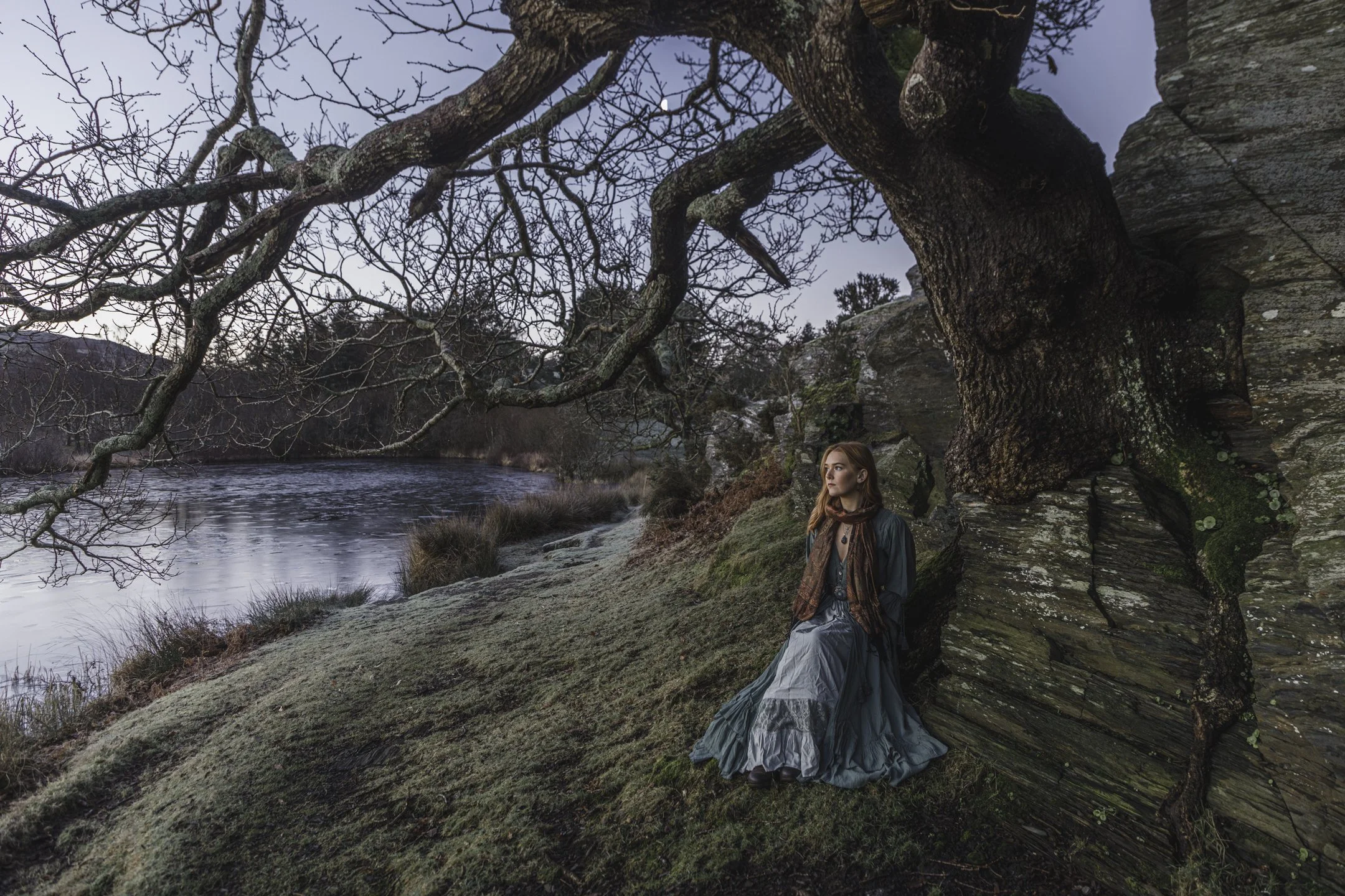 A woman in a long, flowing dress sitting against a large, twisted tree by a river in a natural landscape during dusk or dawn.