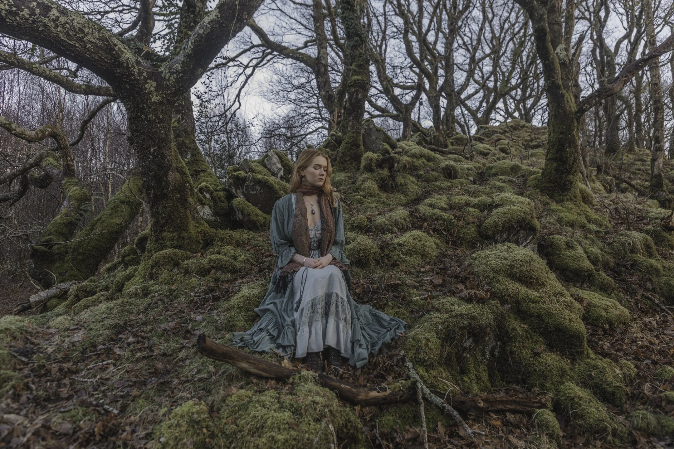 A woman with long, wavy hair sits on a moss-covered forest floor surrounded by leafless trees and rocks, dressed in vintage-style clothing, with a thoughtful expression.