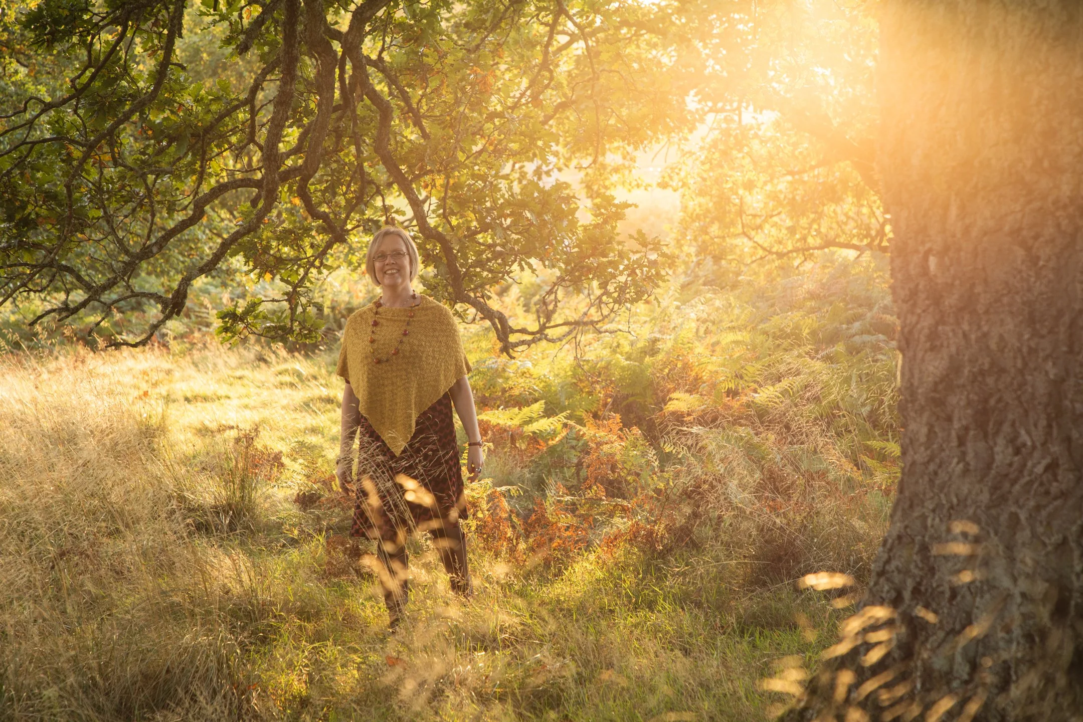 A woman standing in a sunlit forest with trees and foliage around her, wearing a yellow shawl and patterned skirt, smiling