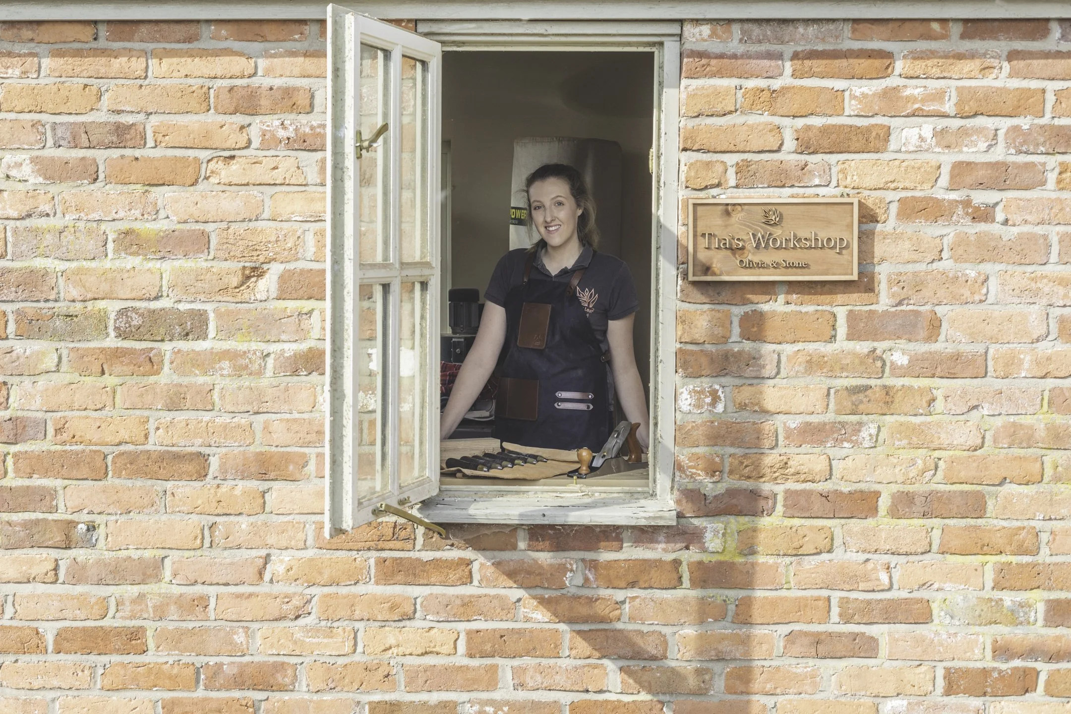 A woman smiling and standing inside a workshop, looking out of an open window. The workshop has a brick exterior and a wooden sign that reads 'Tia's Workshop, Olivia & Stone.' The woman is wearing a dark apron over her clothes, with various tools on 