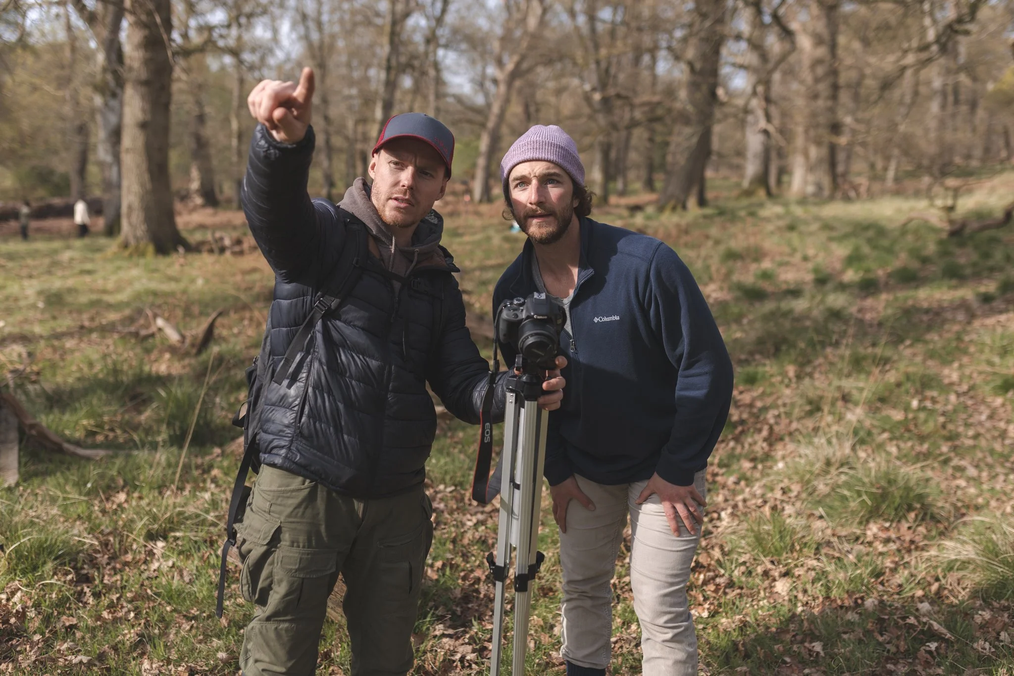 Two men with cameras in a wooded outdoor setting, one pointing and explaining, the other with a camera on a tripod, both dressed in outdoor gear.