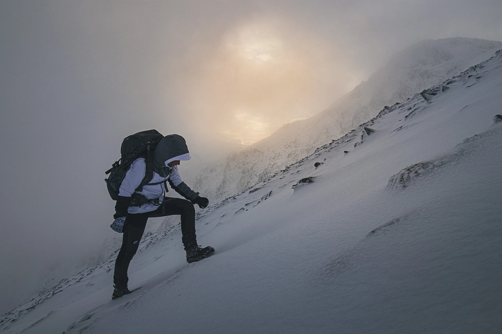 A person hiking up a snowy mountain slope with a backpack, dressed in winter gear, under a cloudy sky.
