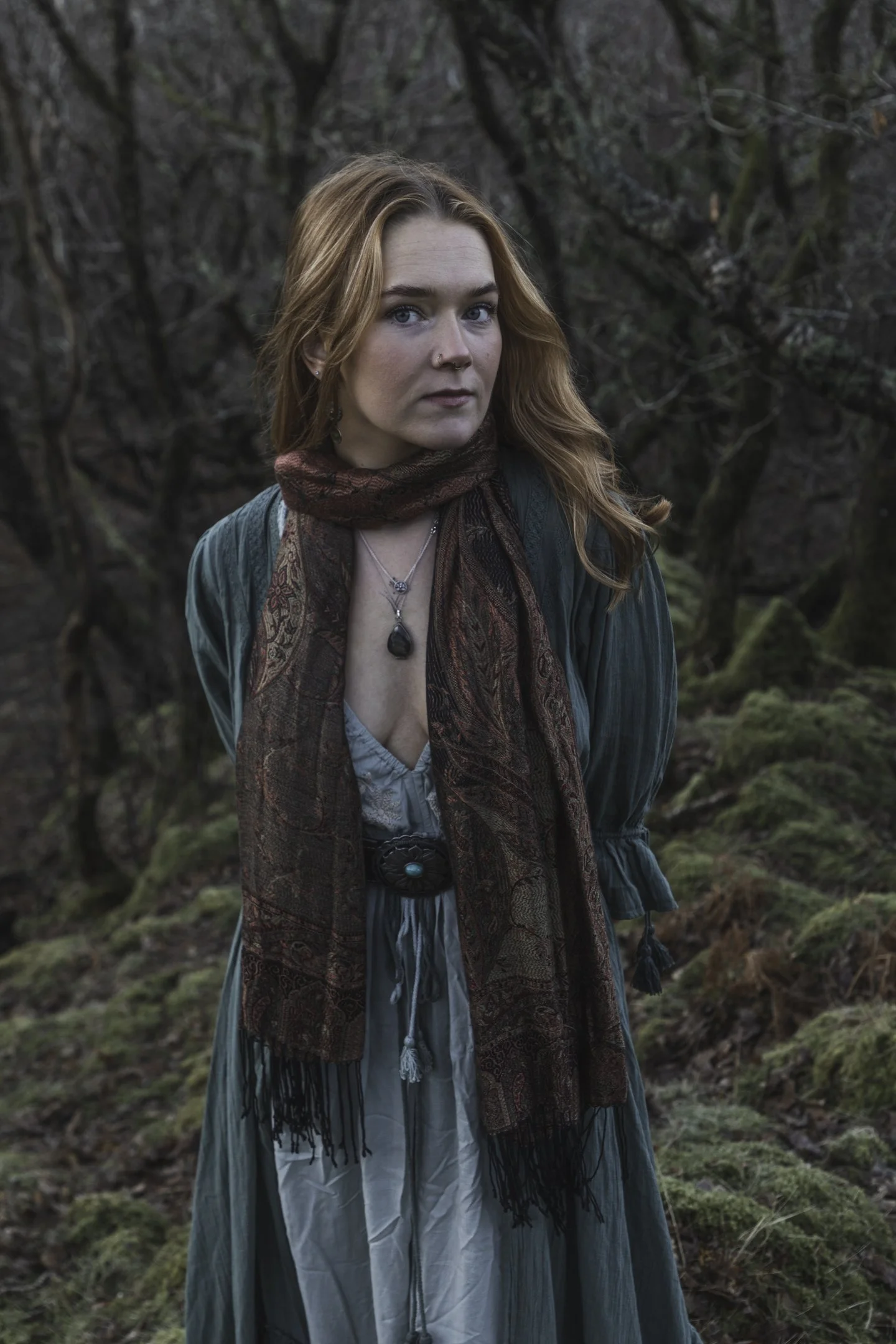 A woman with red hair, blue eyes, and a nose piercing, standing outdoors in a forest with moss-covered ground and leafless trees, wearing a teal dress, a patterned scarf around her neck, and layered necklaces.