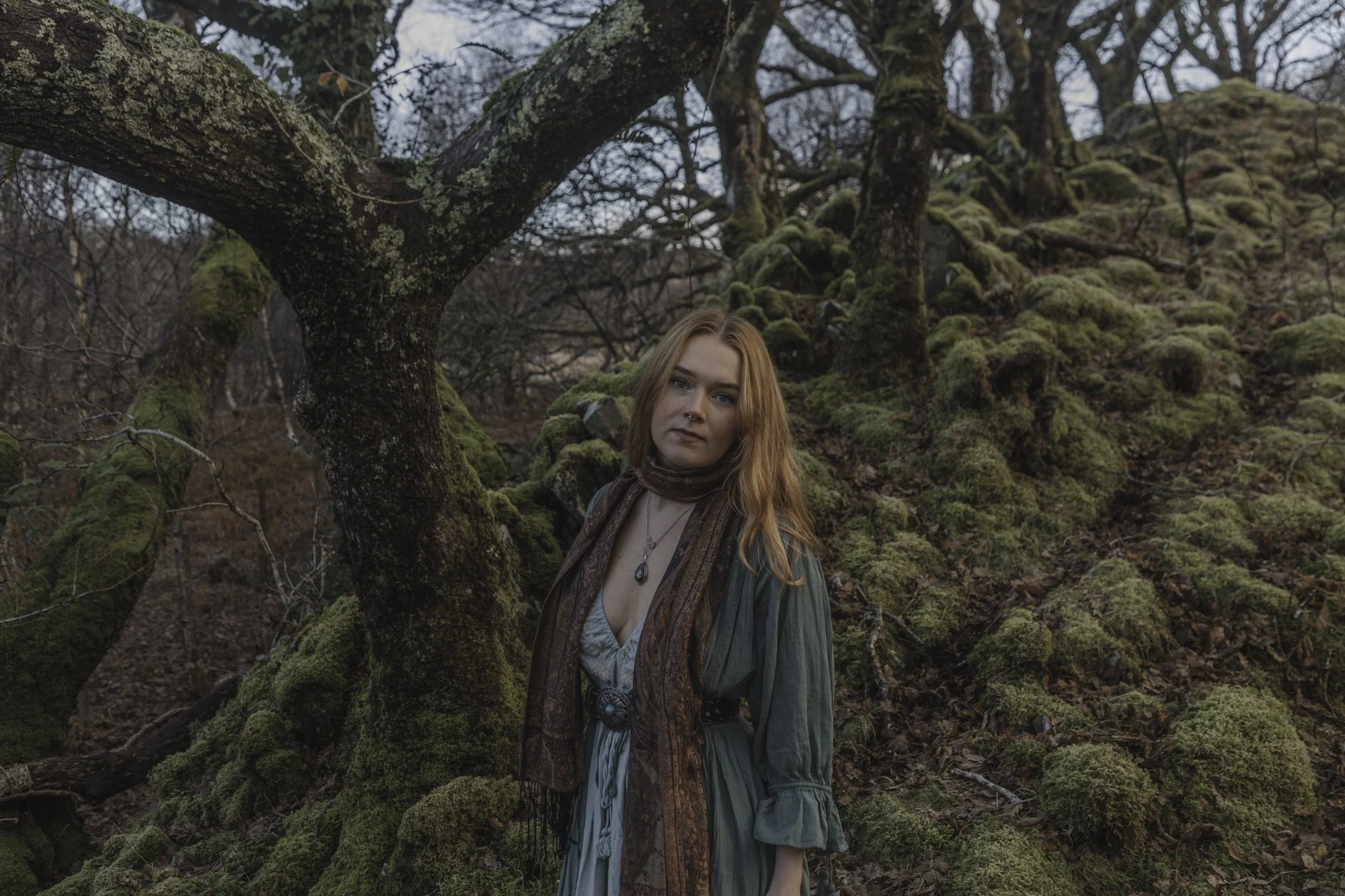 A woman with long red hair standing in a moss-covered forest with leafless trees.